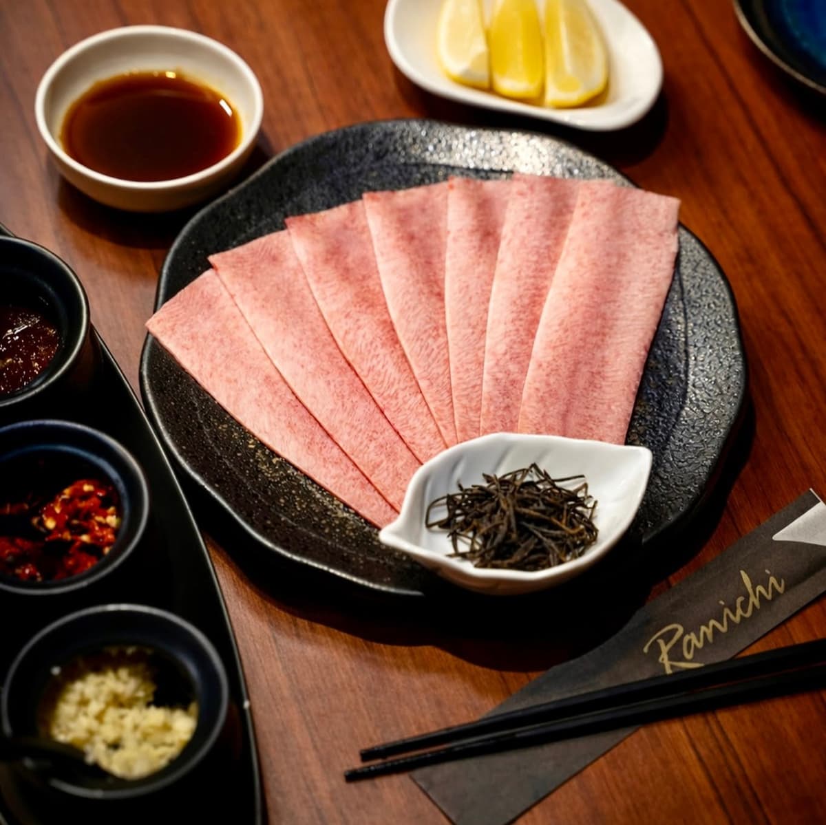 黒い皿に並べられた薄切り肉と薬味、レモン、タレのセット