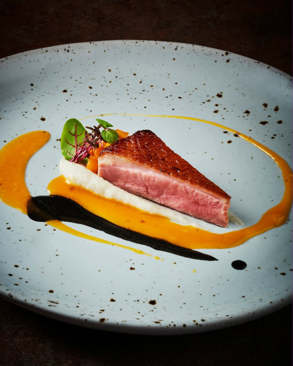 モダンな平皿に美しく盛り付けられた鴨胸肉のロースト。きれいなピンク色の断面に、マンゴーソースと黒いソースがスタイリッシュに描かれている
