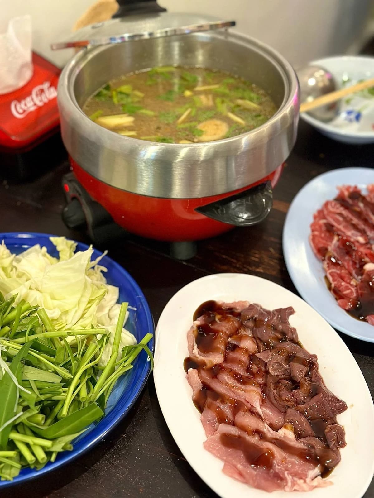 卓上用の赤い電気鍋でグツグツと煮立つタイ風の鍋料理（チムチュム）。手前にはタレがかかった生の豚肉やレバー、そして空芯菜やキャベツなどの新鮮な野菜が青い皿に盛られている