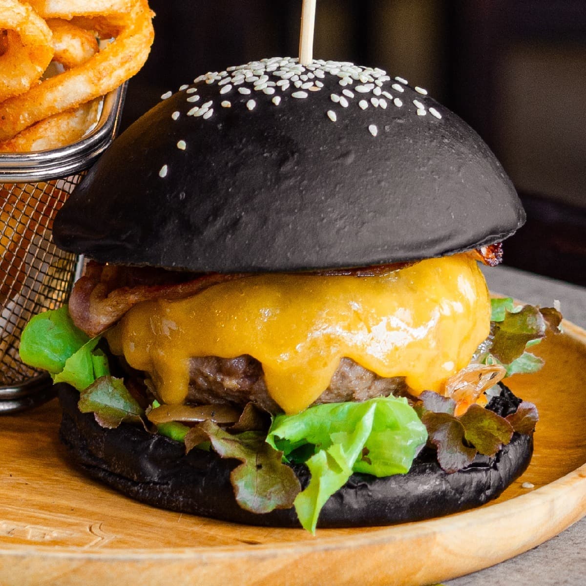 木製のプレートにのせられた、黒いバンズ（炭入りパン）が特徴的なボリュームのあるチーズバーガー。中にはジューシーなパティ、とろけるチーズ、ベーコン、レタスが挟まれており、奥にフレンチフライが添えられている。