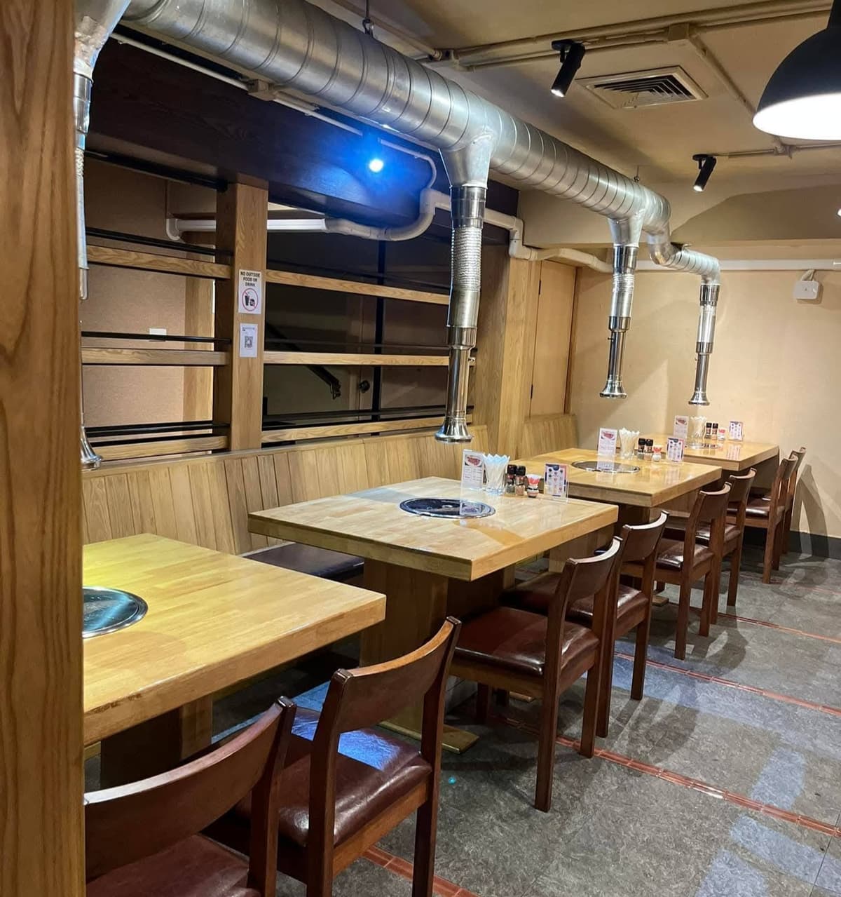 清潔感のある焼肉店の内観。木のテーブルと椅子が並び、各テーブルの上には無煙ロースターと強力な排気ダクトが設置されている。
