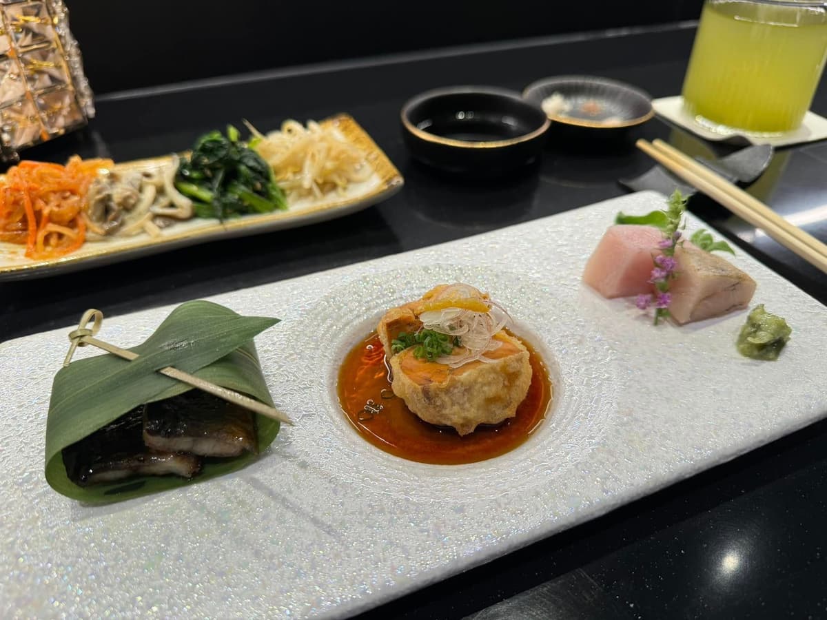 白く細長い長皿に美しく盛り付けられた、笹包みの料理、タレがかかった揚げ物、お刺身などの和食前菜盛り合わせ