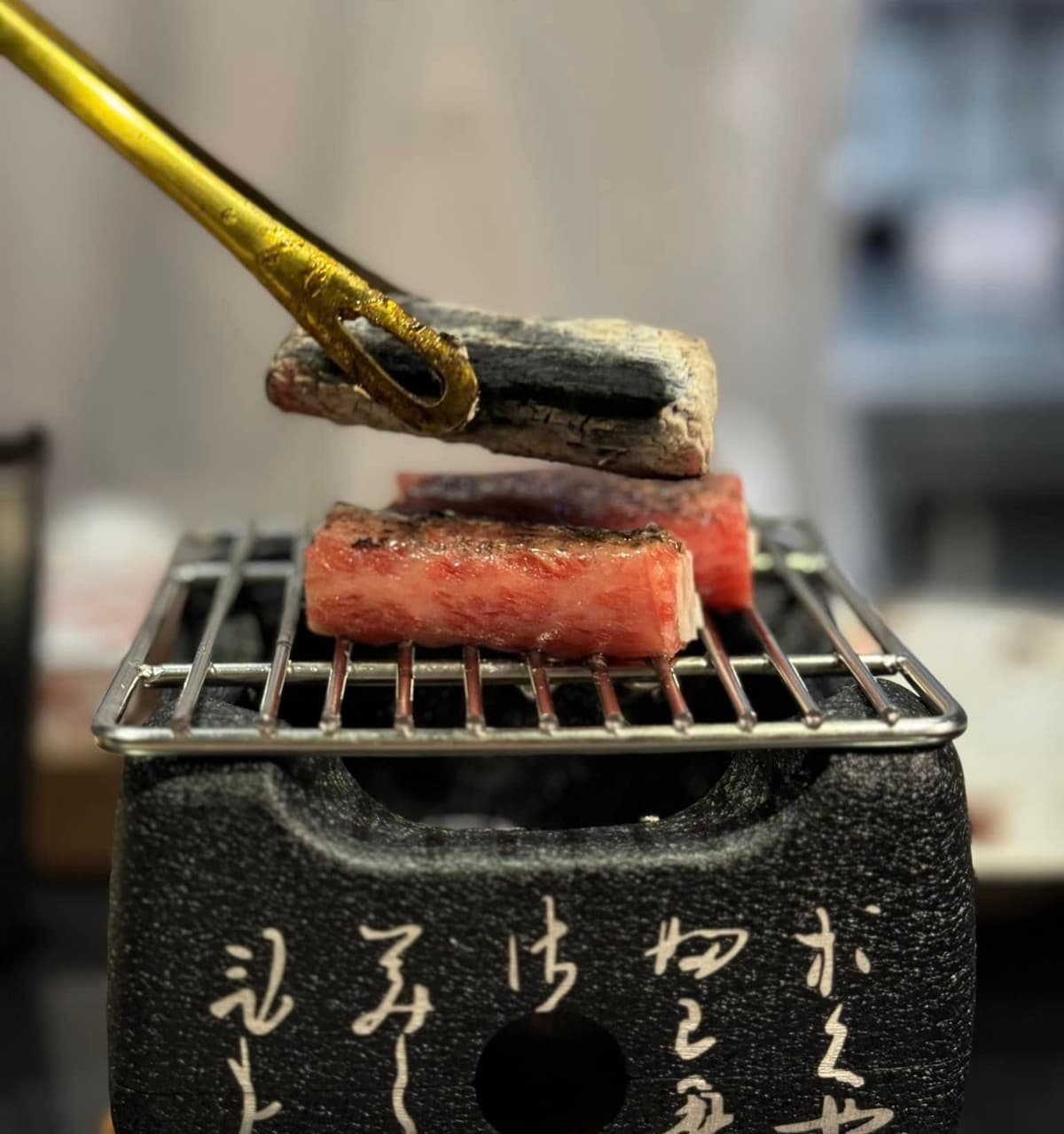 文字が刻まれた小さな卓上七輪の網の上で、トングを使って炭火で焼かれている厚切りの霜降り和牛