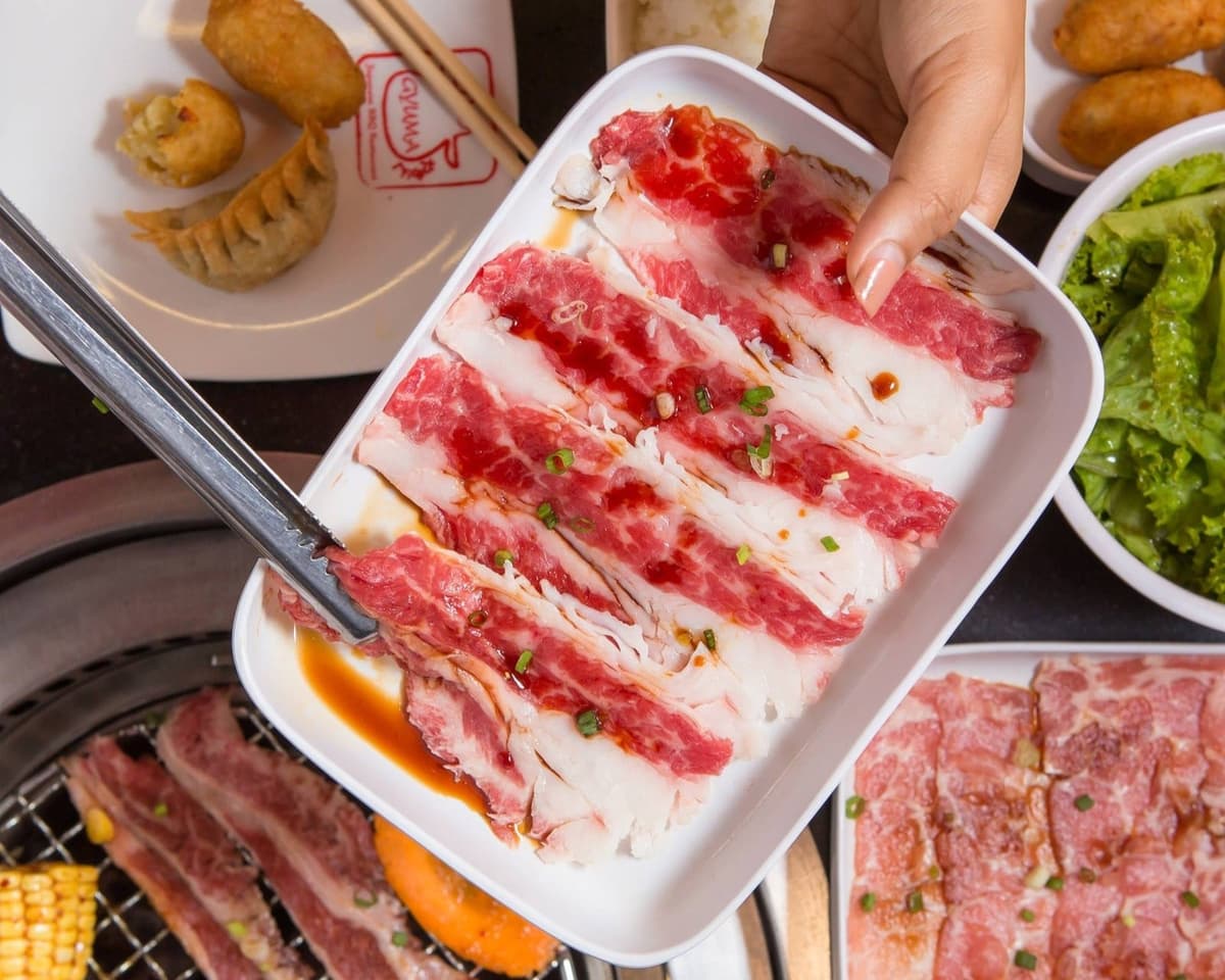 タレをかけた薄切り牛肉の皿を手に持ち、卓上ロースターで焼肉を楽しむ様子