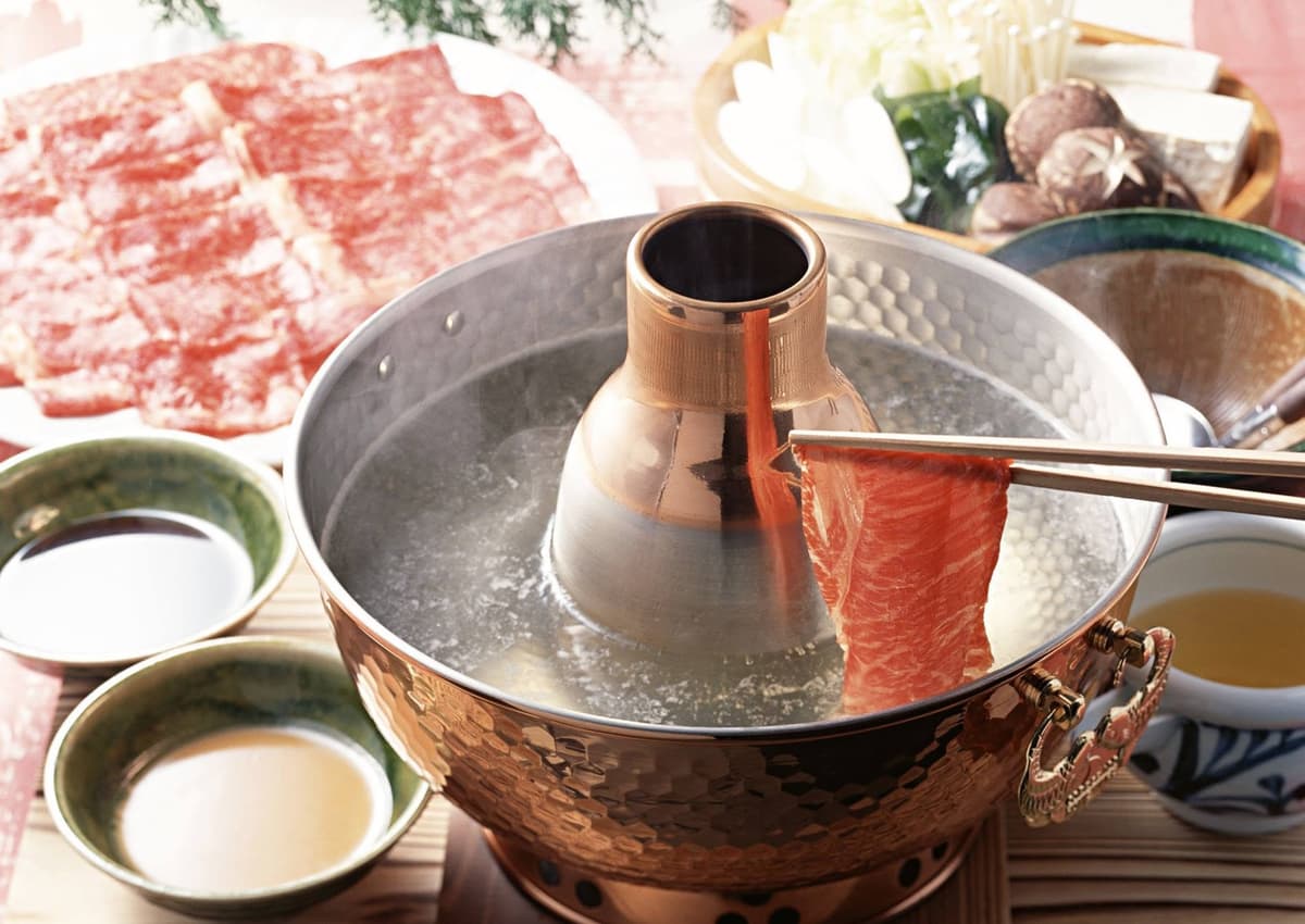 中央に煙突のある伝統的な鍋で、箸で持ち上げた薄切りの牛肉をしゃぶしゃぶしている様子。