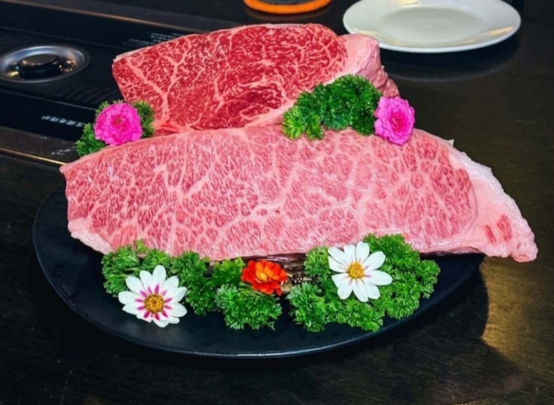 黒い大皿に盛られた美しい霜降りの近江牛の大きな塊肉2つ。周囲にはパセリや鮮やかな花が添えられている。