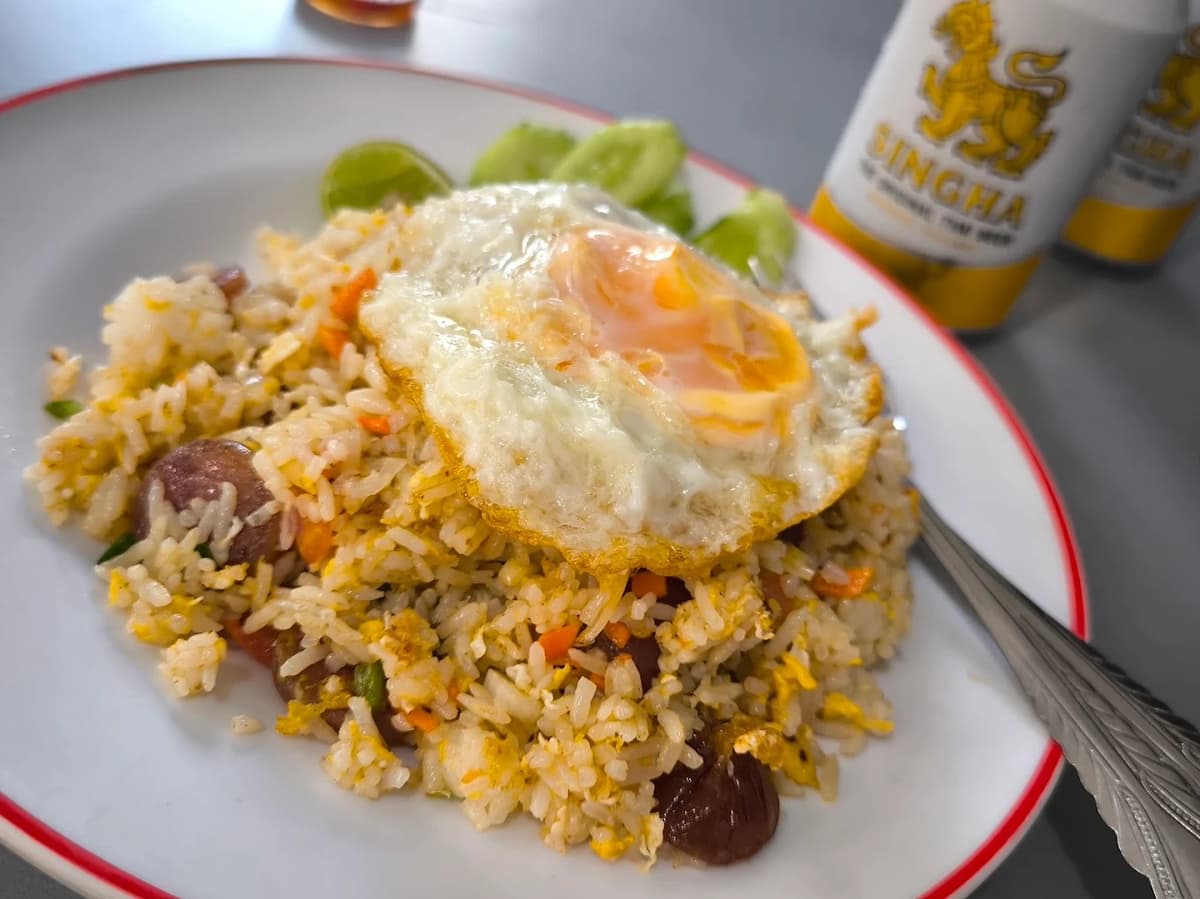 ソーセージ入りのチャーハンに半熟の目玉焼きがのった、シンハービールの缶が奥に見える一皿。