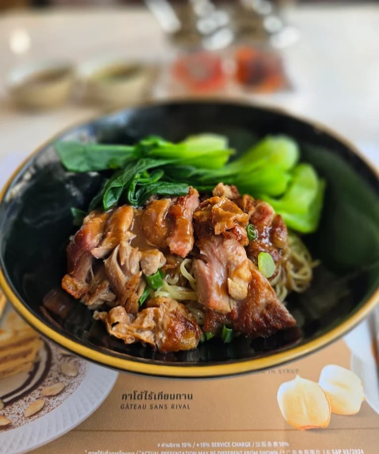 黒いボウルにきれいに盛り付けられた、たっぷりの青菜とジューシーなローストポークがのった卵麺(バミー)料理。