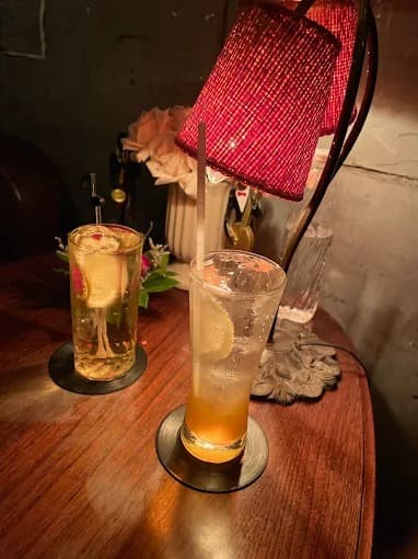 赤い網目状のランプシェードが灯るテーブルの上で、レモンスライスが入った背の高いロンググラスのカクテルと別のグラスドリンクが並んでいる様子。